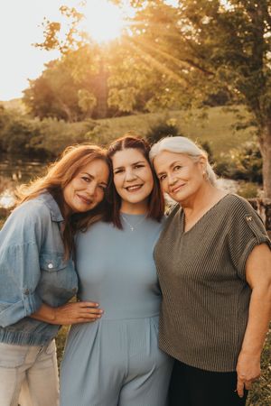 Three women smiling and embracing outdoors during golden hour sunset with lush green trees in background.