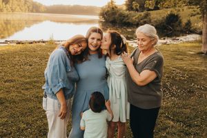 Five women and a child share a warm embrace outdoors near a lake at golden hour in a heartfelt family portrait.