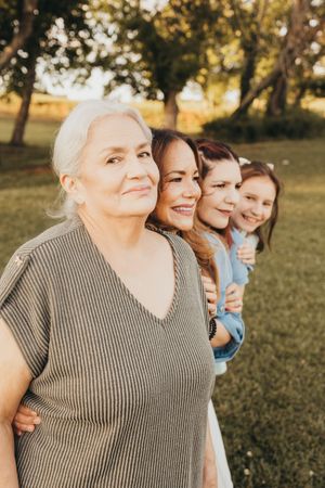 Four generations of women posing in a line outdoors at sunset, smiling warmly in a lush green park setting.