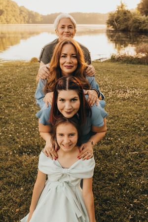 Four generations of women pose together outdoors near a lake at golden hour, smiling warmly at the camera.