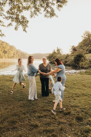 Family gathering by a sunlit river, with adults and children joyfully holding hands on green grass.