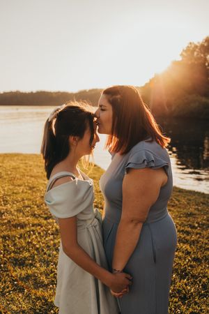 Two women in matching blue dresses share a tender moment at golden hour by a serene lakeside.