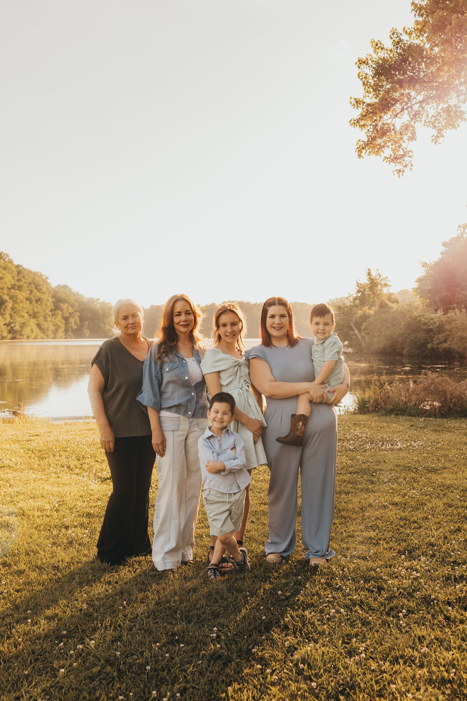 Family group portrait at golden hour by a lake, featuring three women, an older lady, and two young boys on green grass.