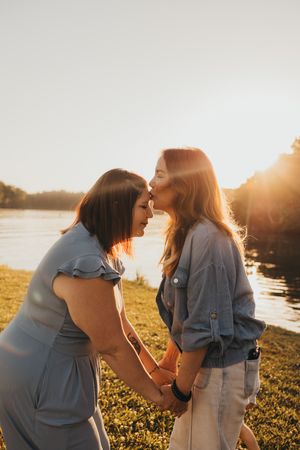 Two women holding hands and sharing a kiss at golden hour by a serene lakeside, bathed in warm sunset light.
