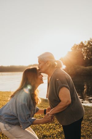 A young woman and elderly lady share a loving kiss at sunset near a peaceful lakeside setting.