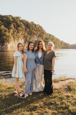 Four women posing together outdoors by a scenic river with tree-covered cliffs at golden hour.
