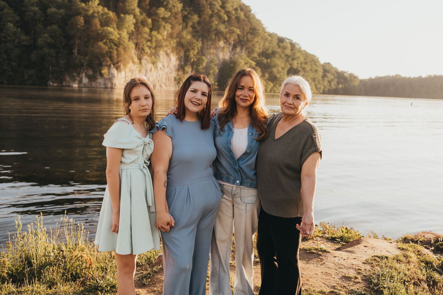 Four women posing together outdoors near a scenic river with rocky cliffs and lush trees in warm golden sunlight.