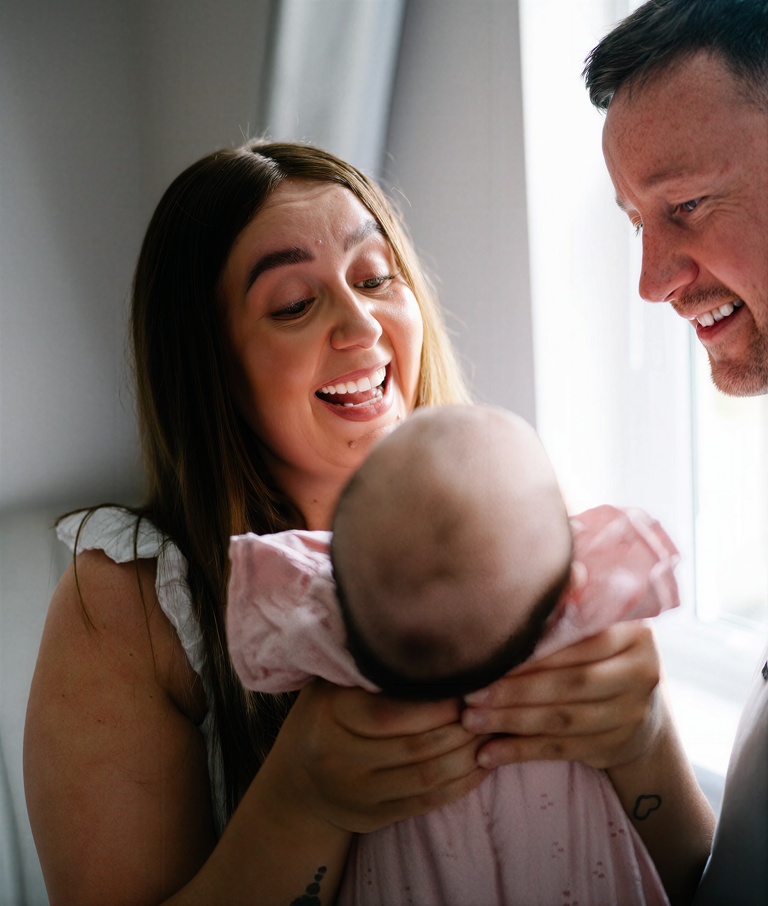 Happy parents smiling and laughing while holding their newborn baby dressed in pink near a bright window.