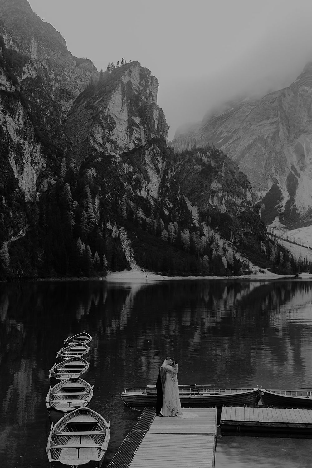 Black and white photo of a couple embracing on a dock by a mountain lake with rowboats and misty peaks reflected in calm water.
