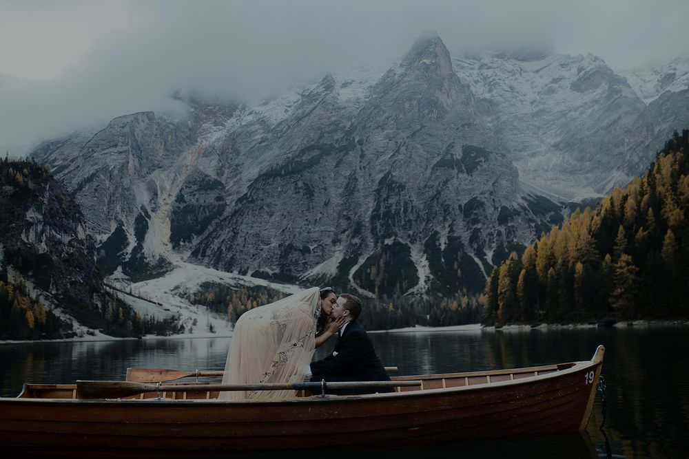 Couple sharing a romantic kiss in a wooden boat on a serene alpine lake with snow-capped Dolomite mountains backdrop.