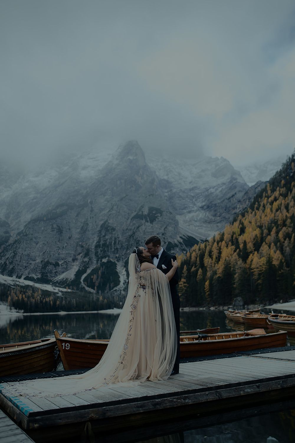 Bride and groom kissing on a dock at Lago di Braies, with snow-capped mountains and autumn trees in the background.