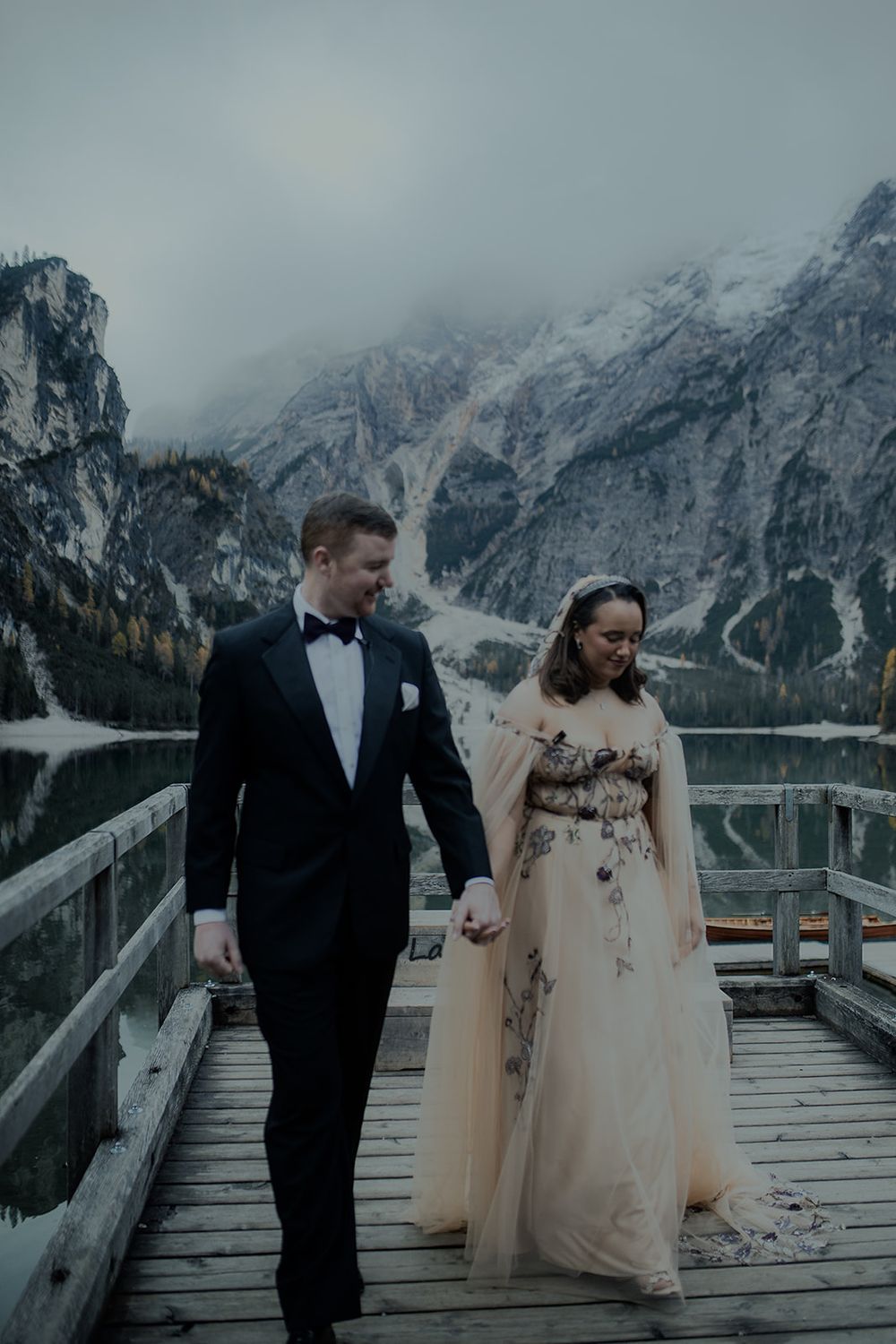 Couple walks hand-in-hand on a dock, bride in floral gown, groom in tuxedo, misty alpine lake backdrop.