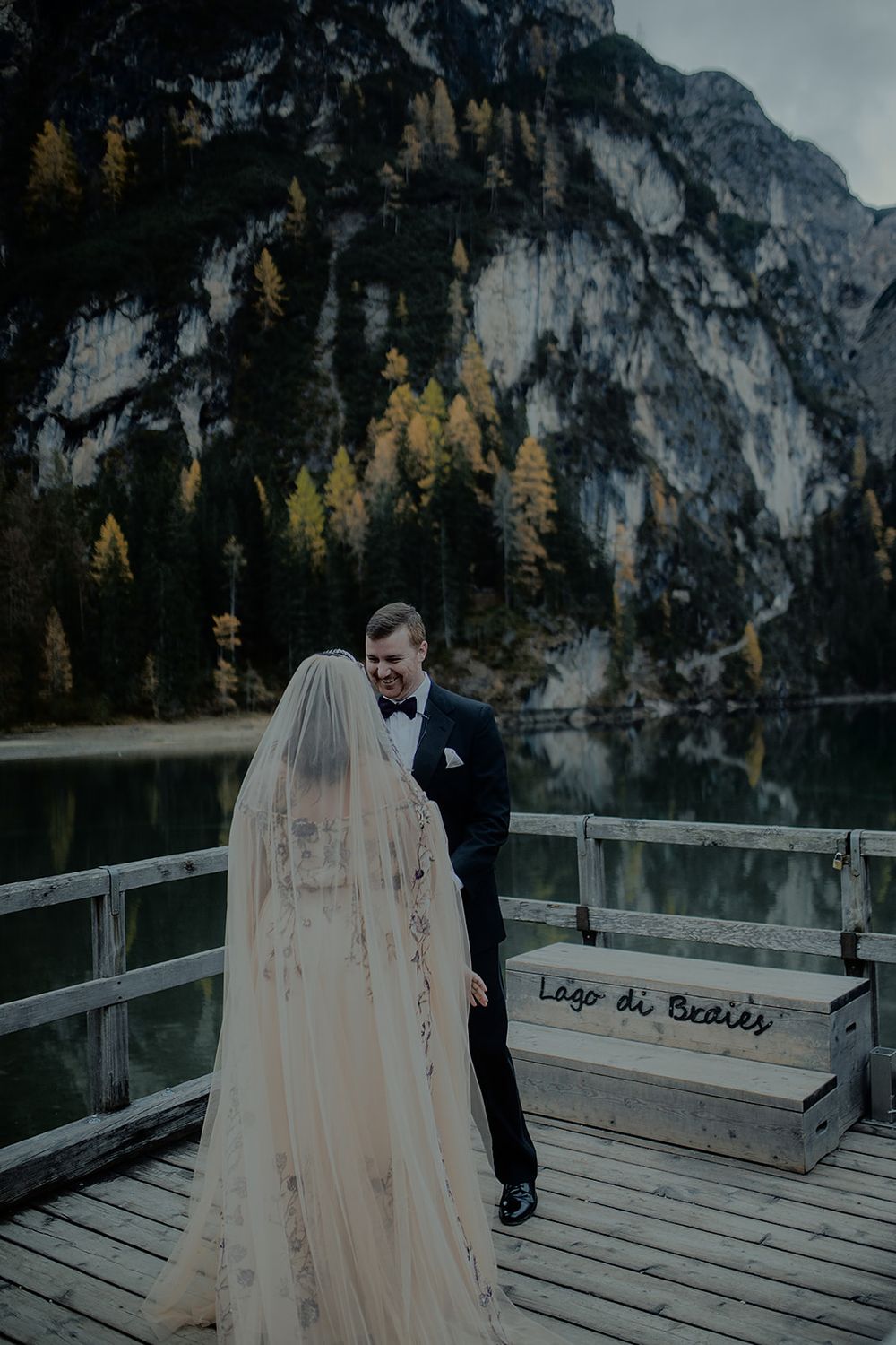 Bride and groom share a romantic moment on a wooden dock at Lago di Braies, Italy, with mountain scenery.