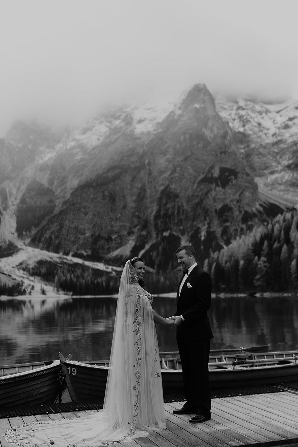 Black and white photo of bride and groom holding hands on a dock with misty mountain lake behind them.