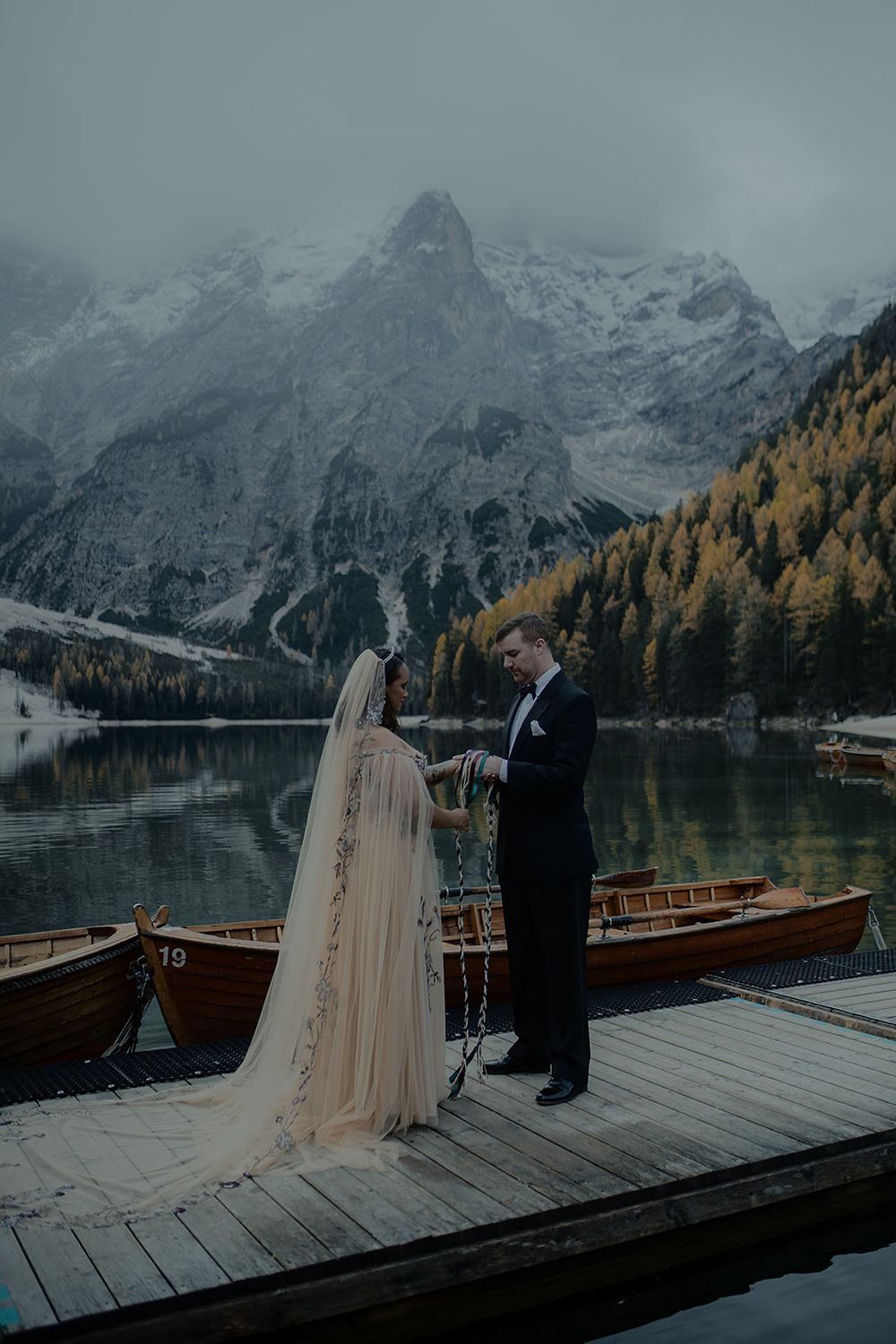 Bride in flowing veil and groom exchange vows on dock at Lago di Braies with misty mountains and autumn trees behind.