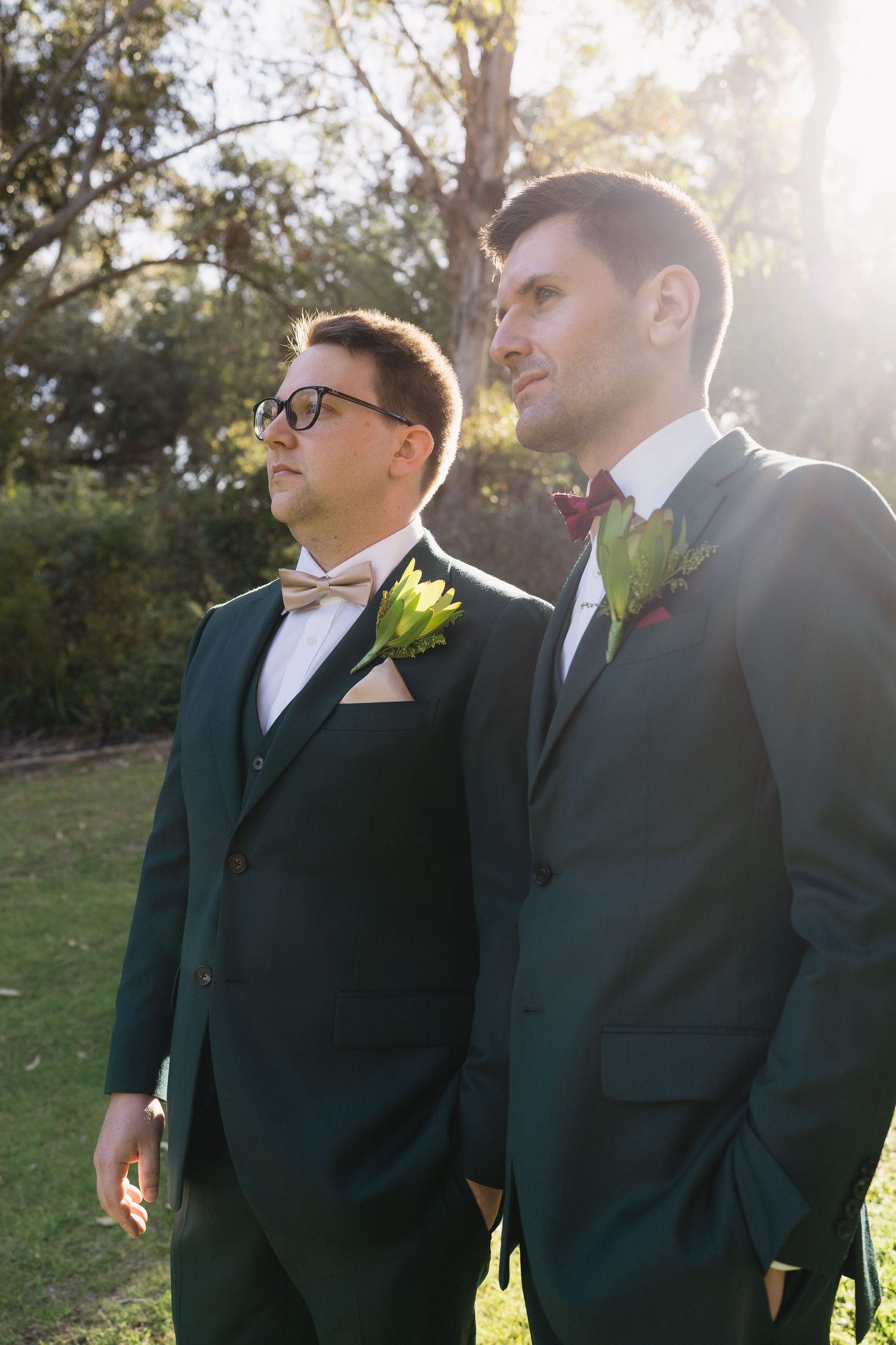 Two grooms in dark green suits with boutonnieres stand outdoors in sunlight at their wedding ceremony.