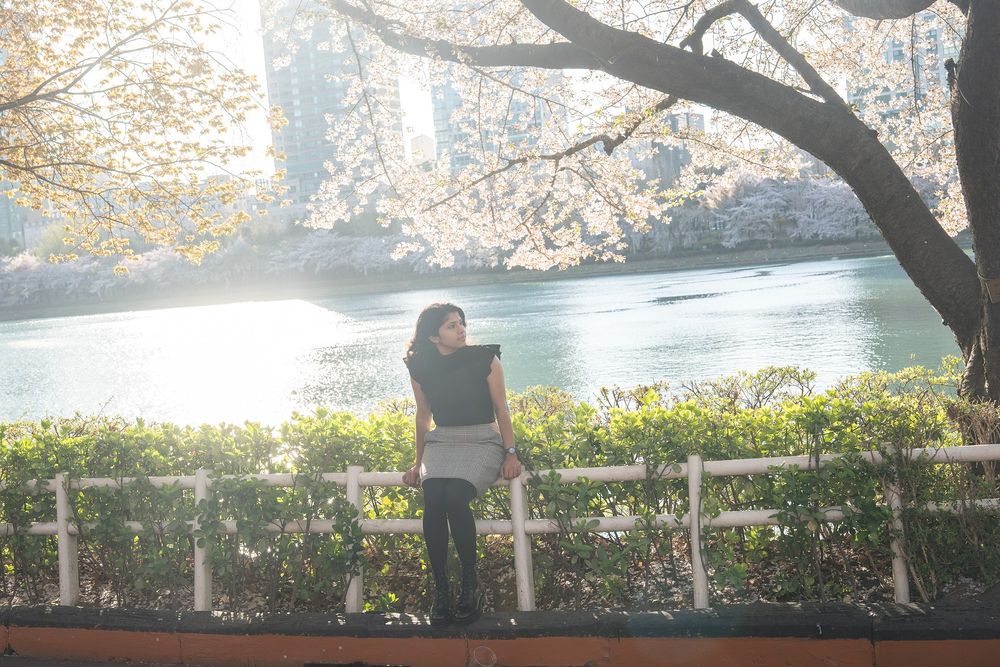 Woman standing by a white fence near a serene river, framed by cherry blossom trees with city buildings in background.