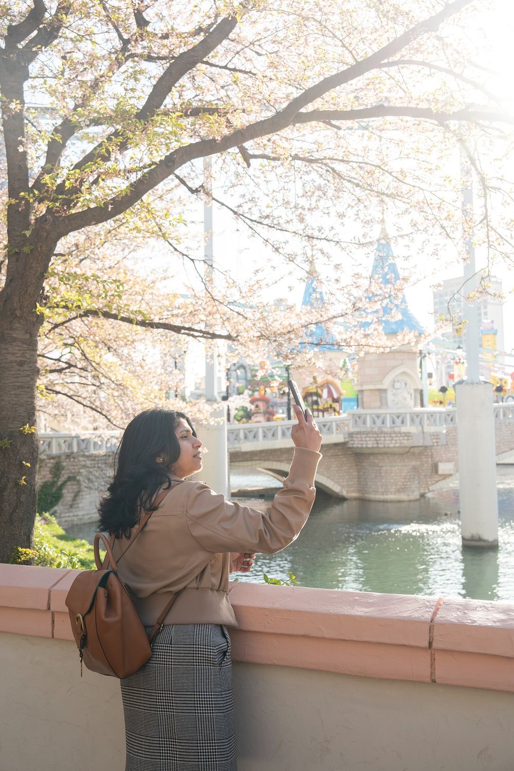 Woman taking a photo with smartphone under cherry blossom trees near a scenic canal in spring.