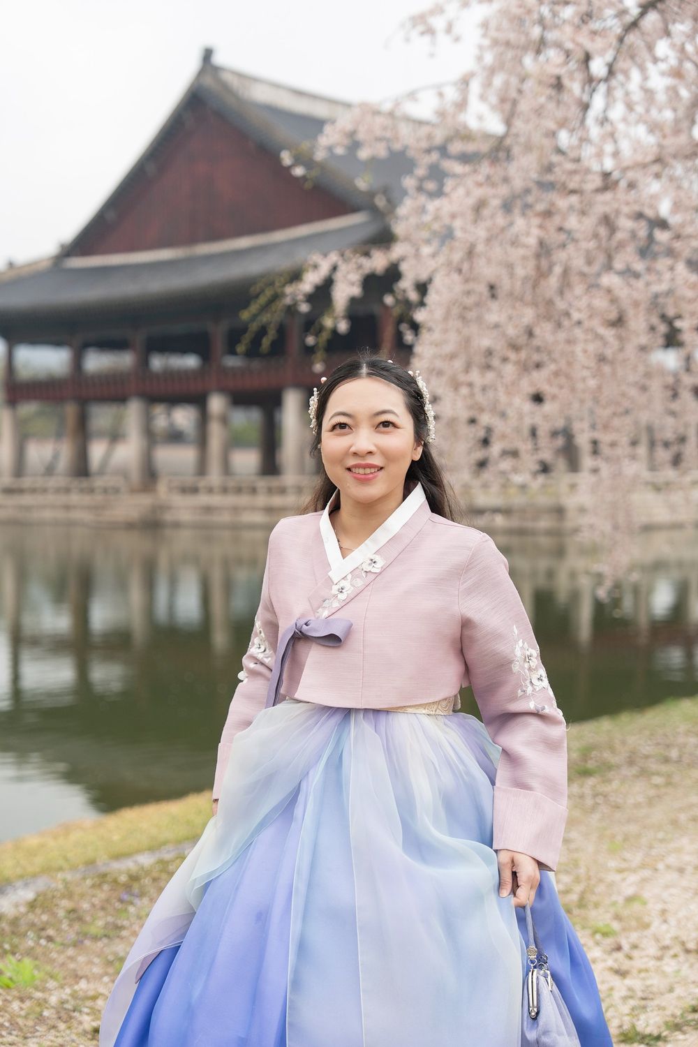 Woman in traditional Korean hanbok dress poses near a pond with cherry blossoms and historic pavilion.