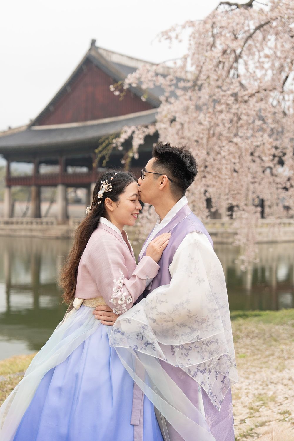 Couple in traditional Korean hanbok embracing near a pond with cherry blossoms and historic pavilion.