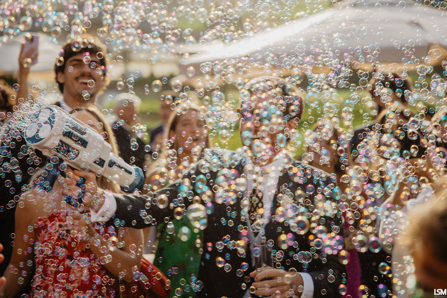 Joyful wedding celebration with guests surrounded by colorful soap bubbles, bride and groom laughing outdoors.