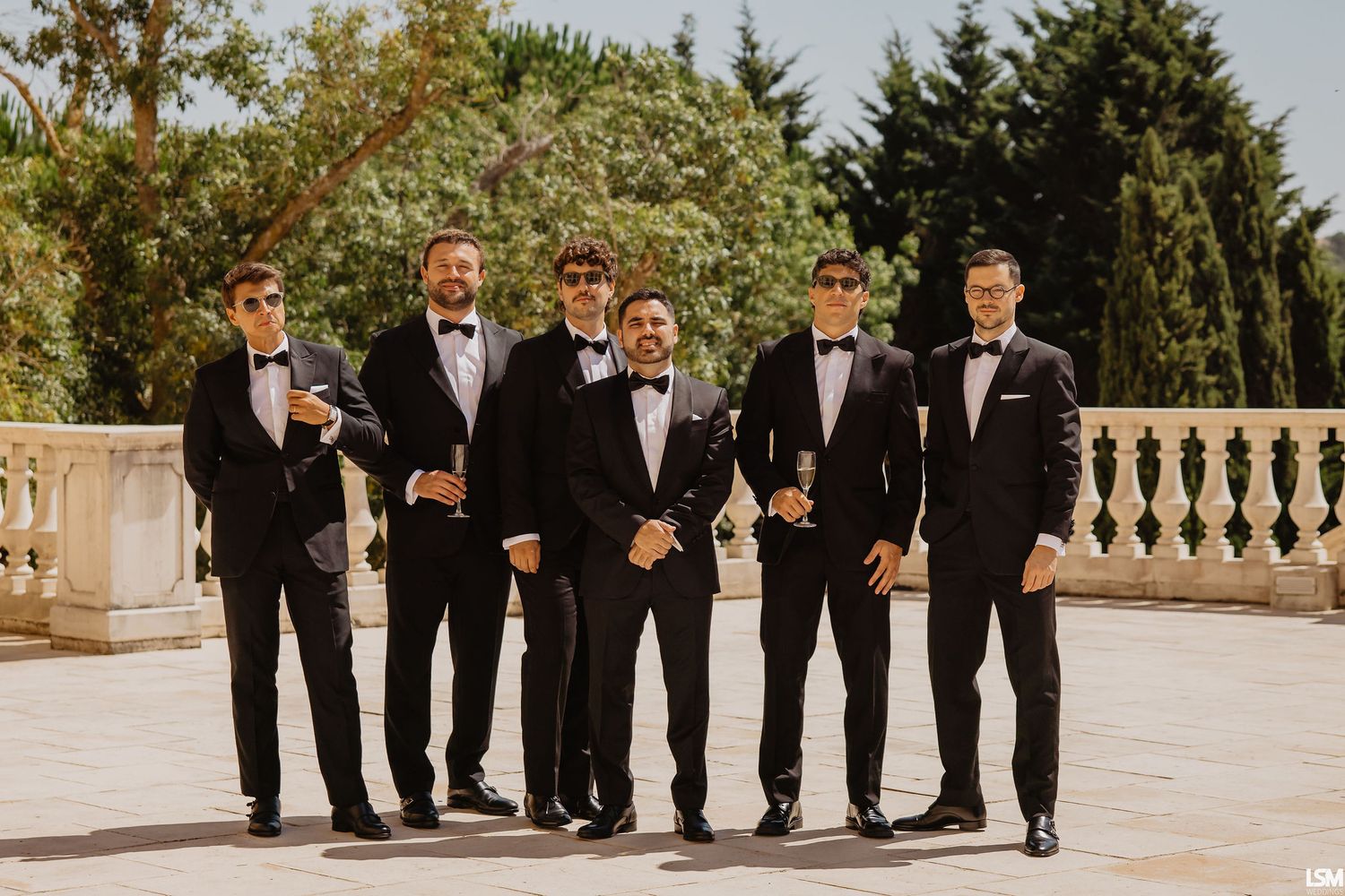 Six groomsmen in black tuxedos and bow ties posing outdoors on a stone terrace at a wedding celebration.