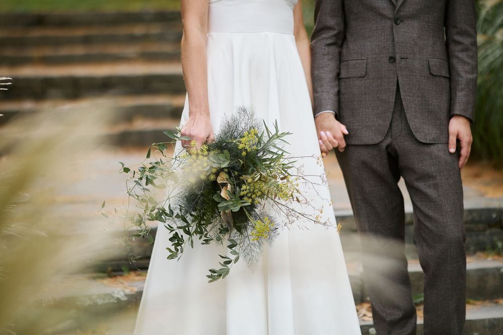 Bride in white gown holding greenery bouquet stands beside groom in gray suit on stone steps.