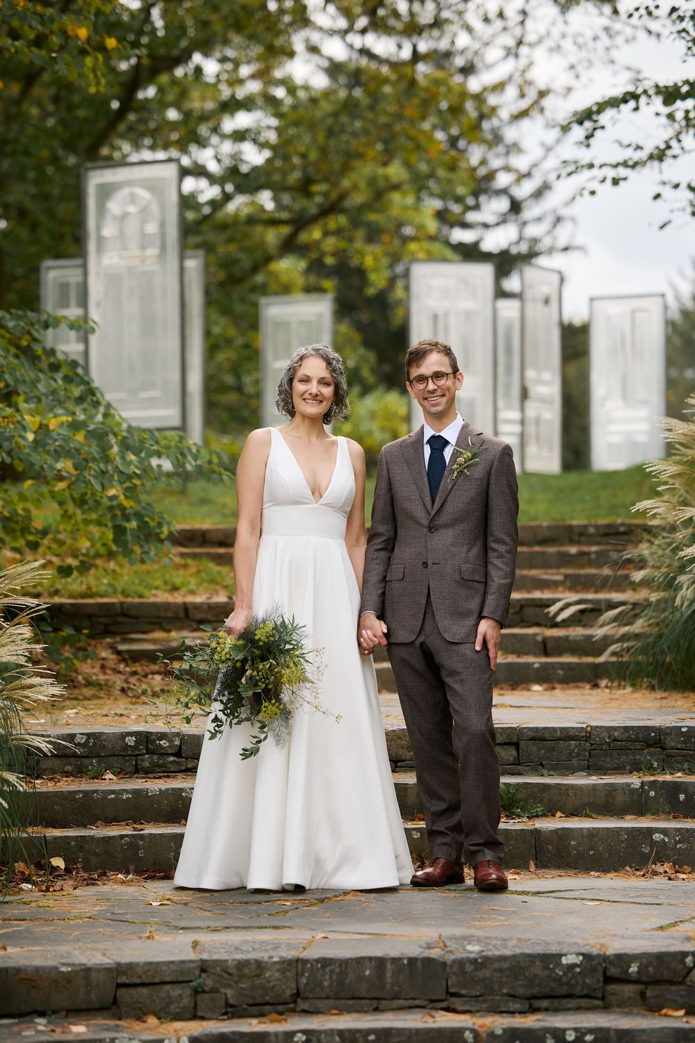 A bride and groom pose on stone steps in a lush garden, she in a white gown holding greenery, he in a gray suit.