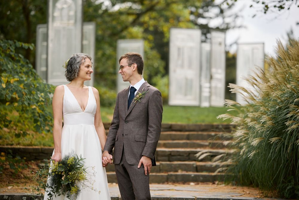 Bride and groom hold hands smiling at each other outdoors in autumn garden setting, bride in white gown holding greenery bouquet.