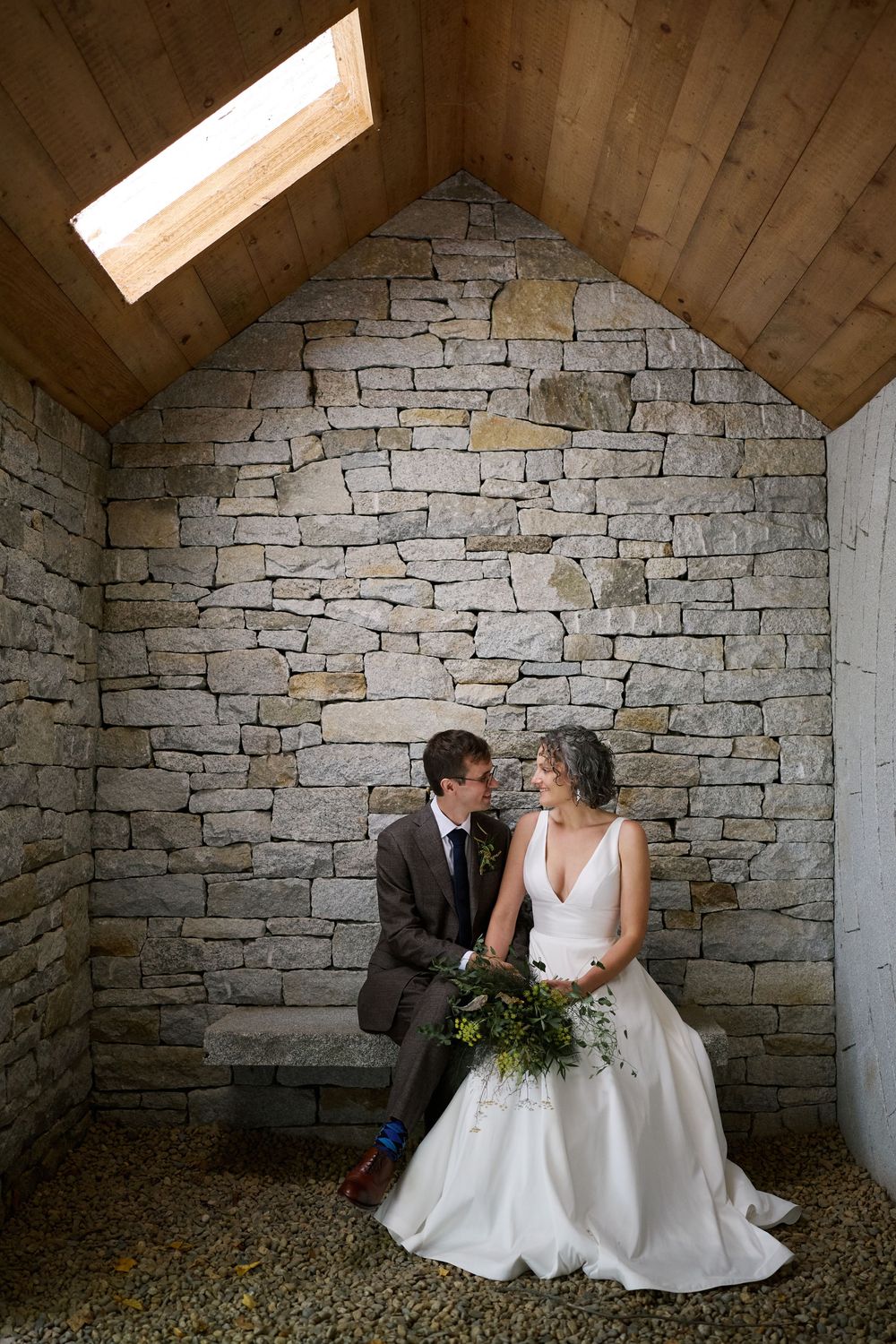Bride and groom sit together against a stone wall under a skylight in a rustic chapel, sharing an intimate moment.