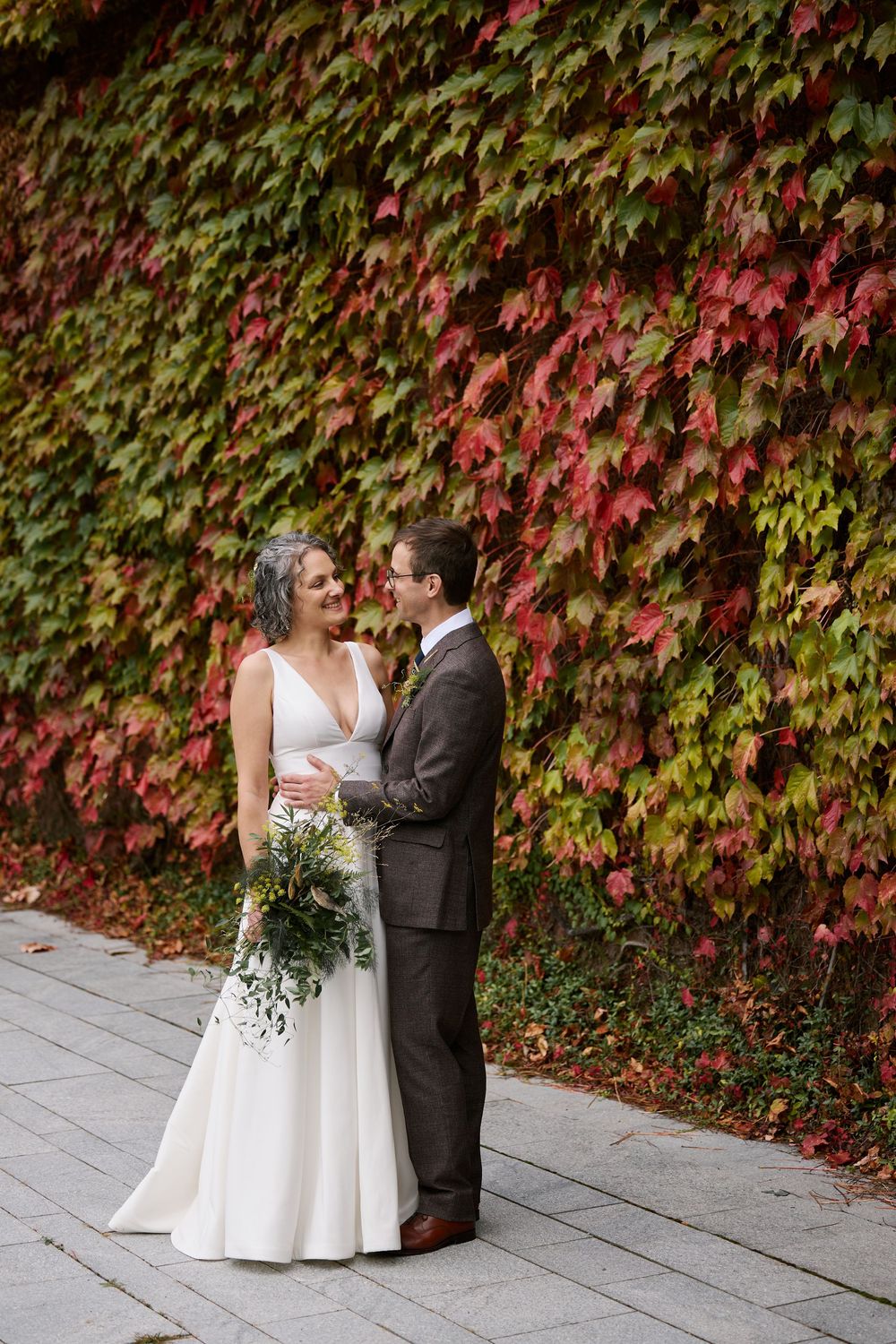 Bride and groom embrace before a stunning autumn ivy wall with red and green leaves on their wedding day.