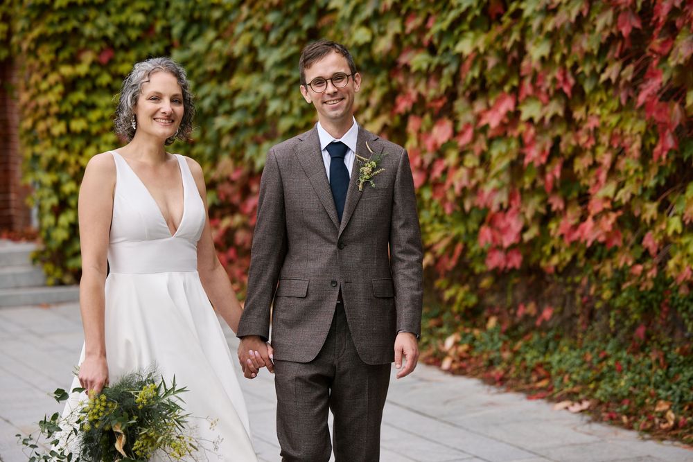 Smiling bride and groom holding hands in front of a vibrant red ivy wall in autumn.