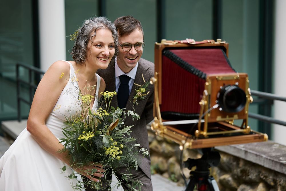Bride and groom smiling beside a vintage large format camera outdoors on their wedding day.