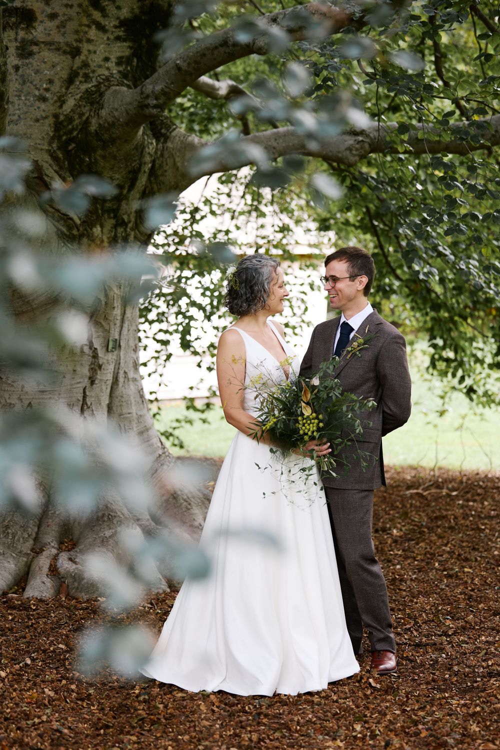 Bride and groom stand under a large tree on wood chip ground, bride holds green bouquet, groom wears brown suit.