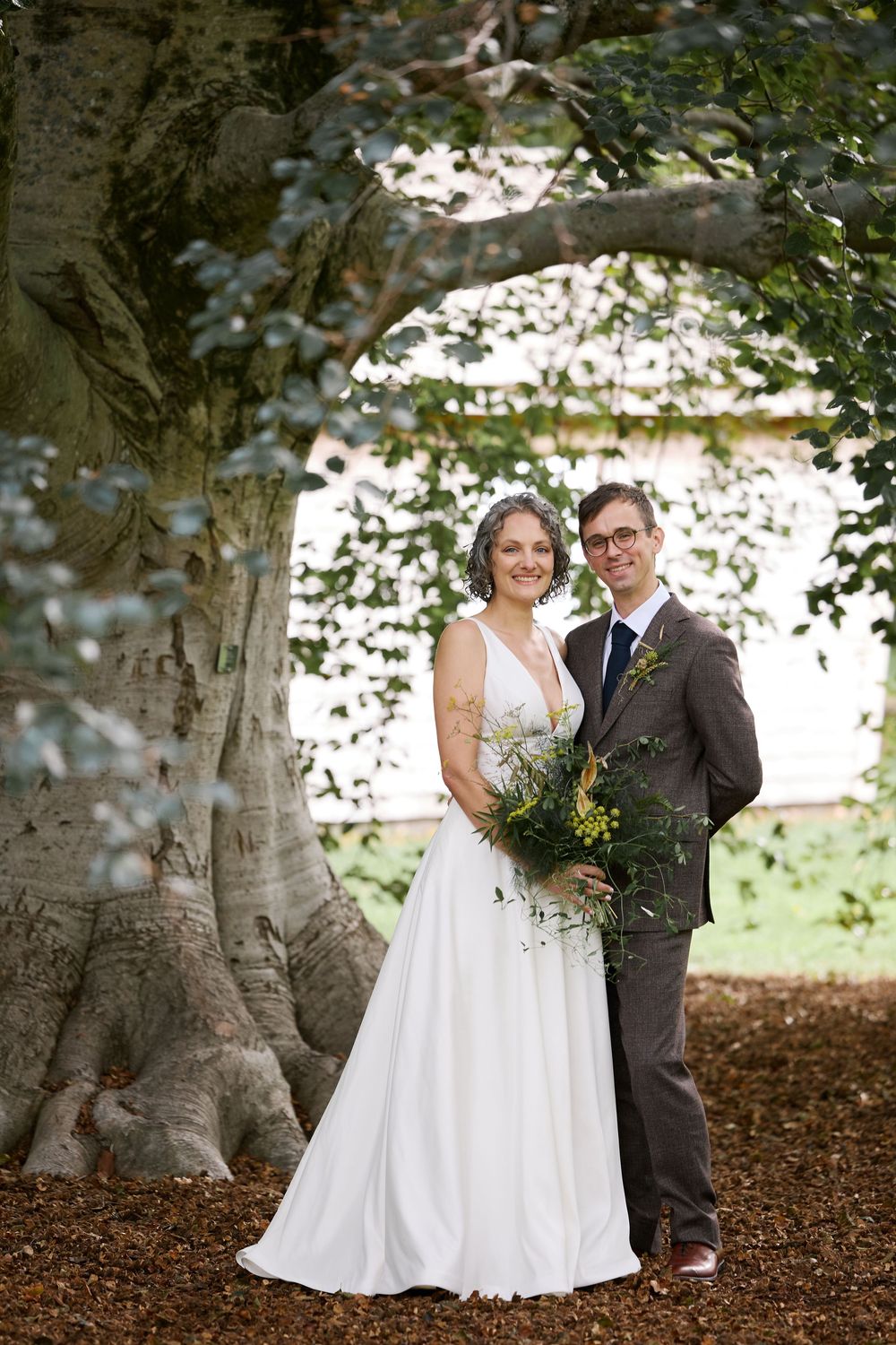 Bride and groom pose together under a large tree, bride holding a wildflower bouquet in a white gown.