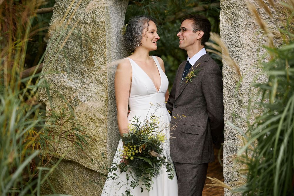 Bride holding green bouquet and groom in brown suit share a romantic moment among stone pillars outdoors.
