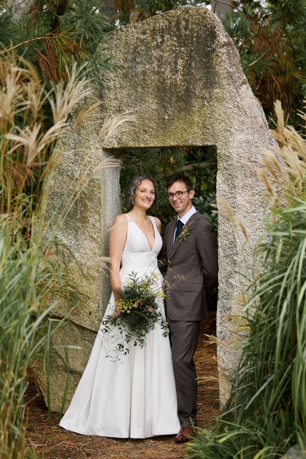 Bride and groom pose in a stone archway surrounded by lush greenery, bride holding a bouquet of flowers.