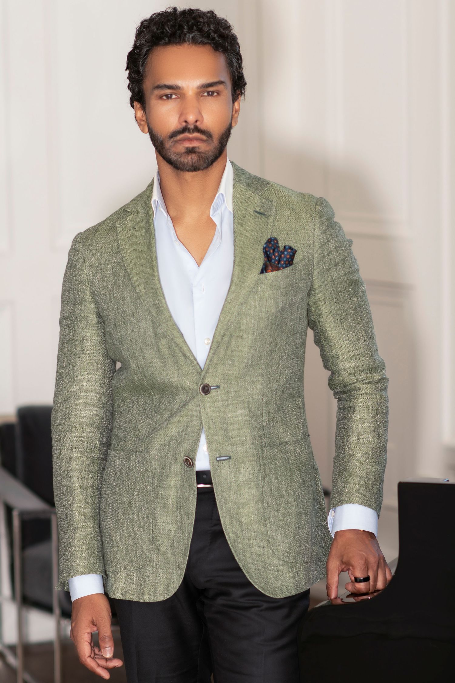 Man in green linen blazer with pocket square, white shirt, and dark trousers posing in elegant interior.
