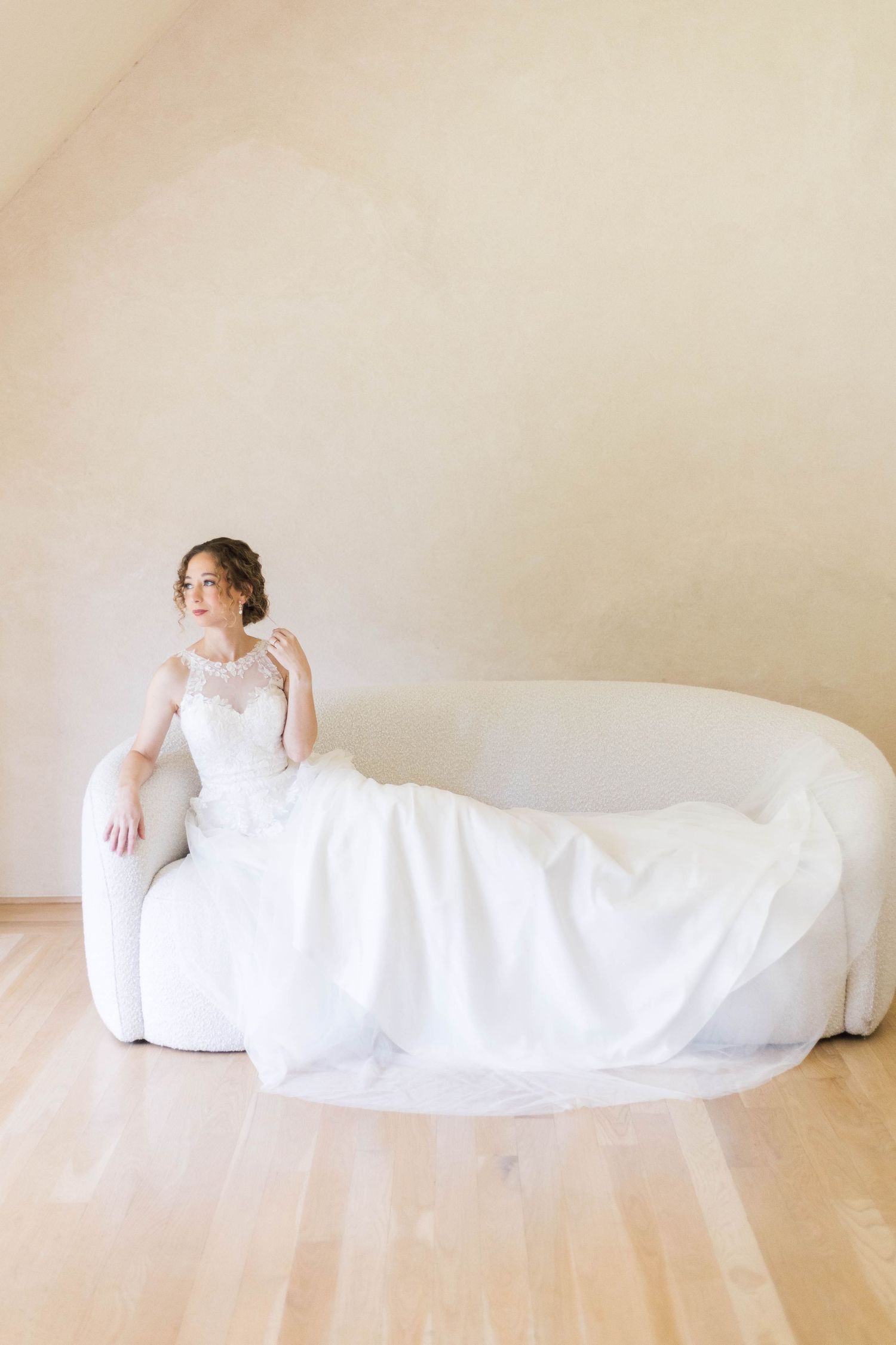 Elegant bride in white lace gown seated gracefully on a cream sofa in a softly lit minimalist room.