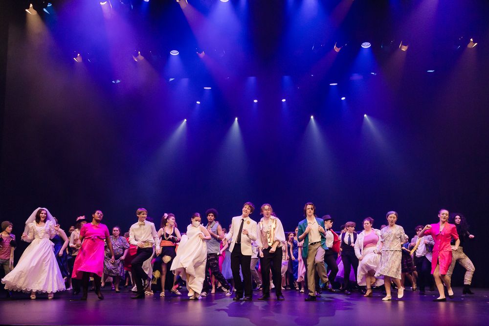 Large cast of performers take a bow on a colorfully lit stage during a theatrical production finale.