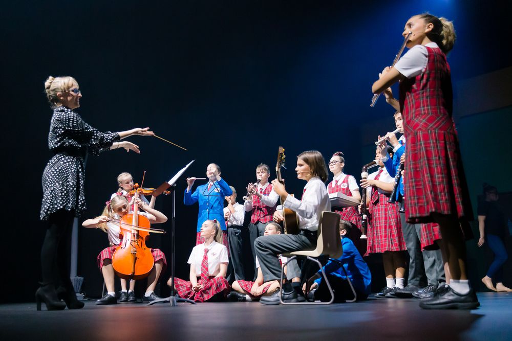 School conductor leads young student orchestra and choir performers on a dark stage.