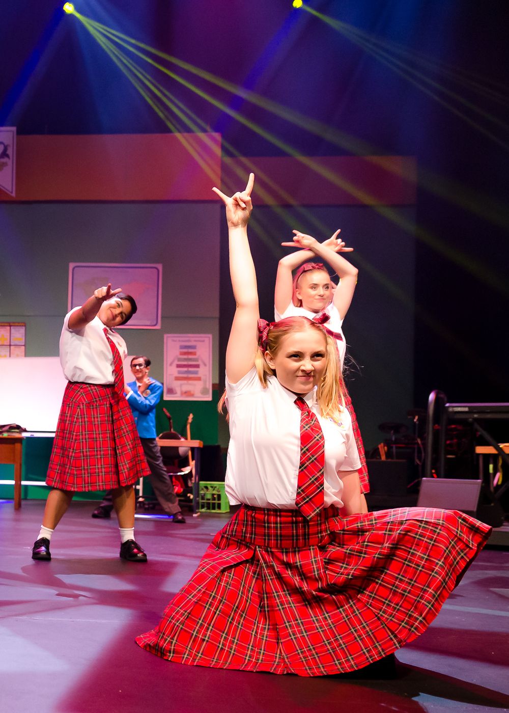 Students in red tartan uniforms dance energetically under colorful stage lights in school musical.