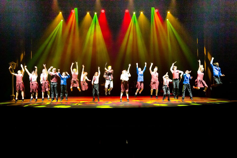Large cast of performers take a bow on stage under vibrant red and green stage lighting.