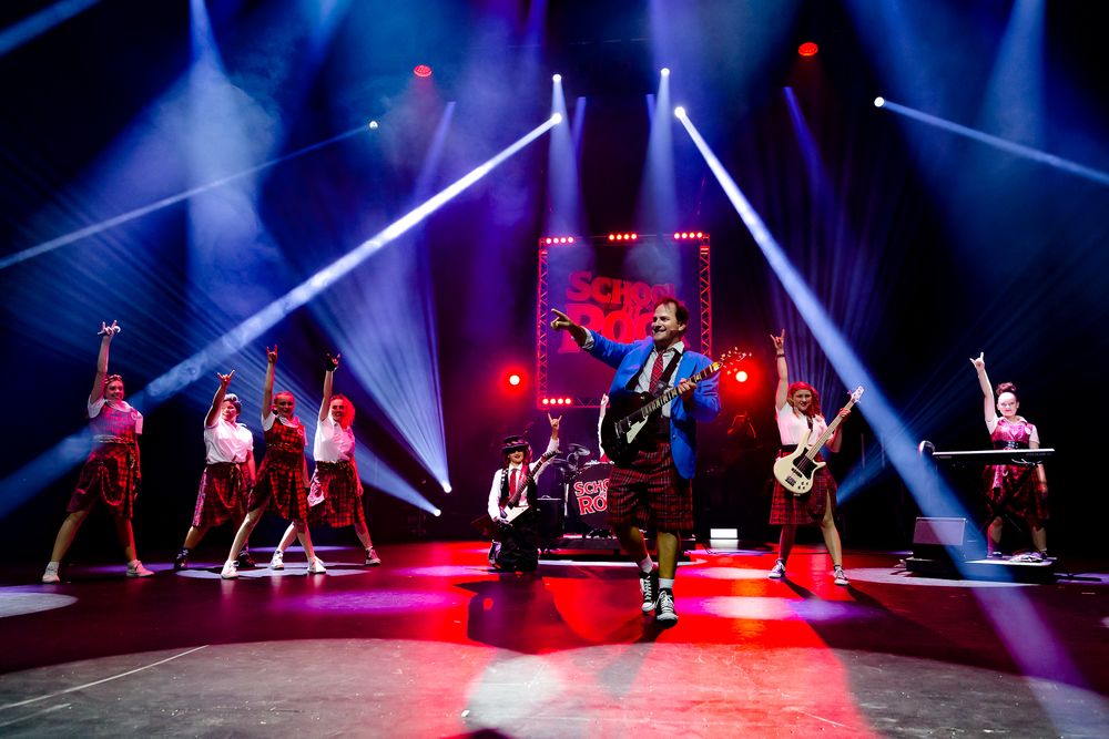 Energetic school rock musical finale with cast in tartan uniforms under dramatic stage lighting.
