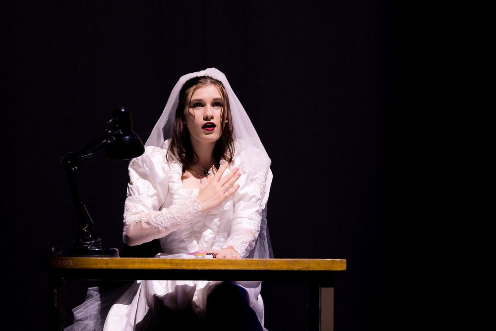 Young performer dressed as a bride sits at a desk on a darkened theater stage during a production.