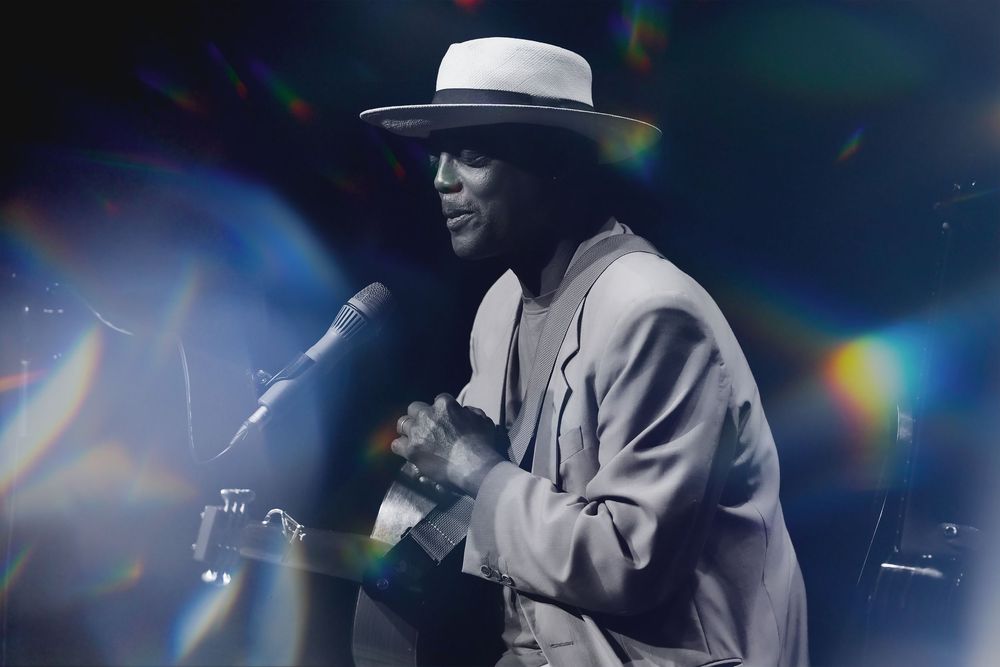 Male performer in a white suit and hat sings into a microphone on a dramatic, light-filled stage.