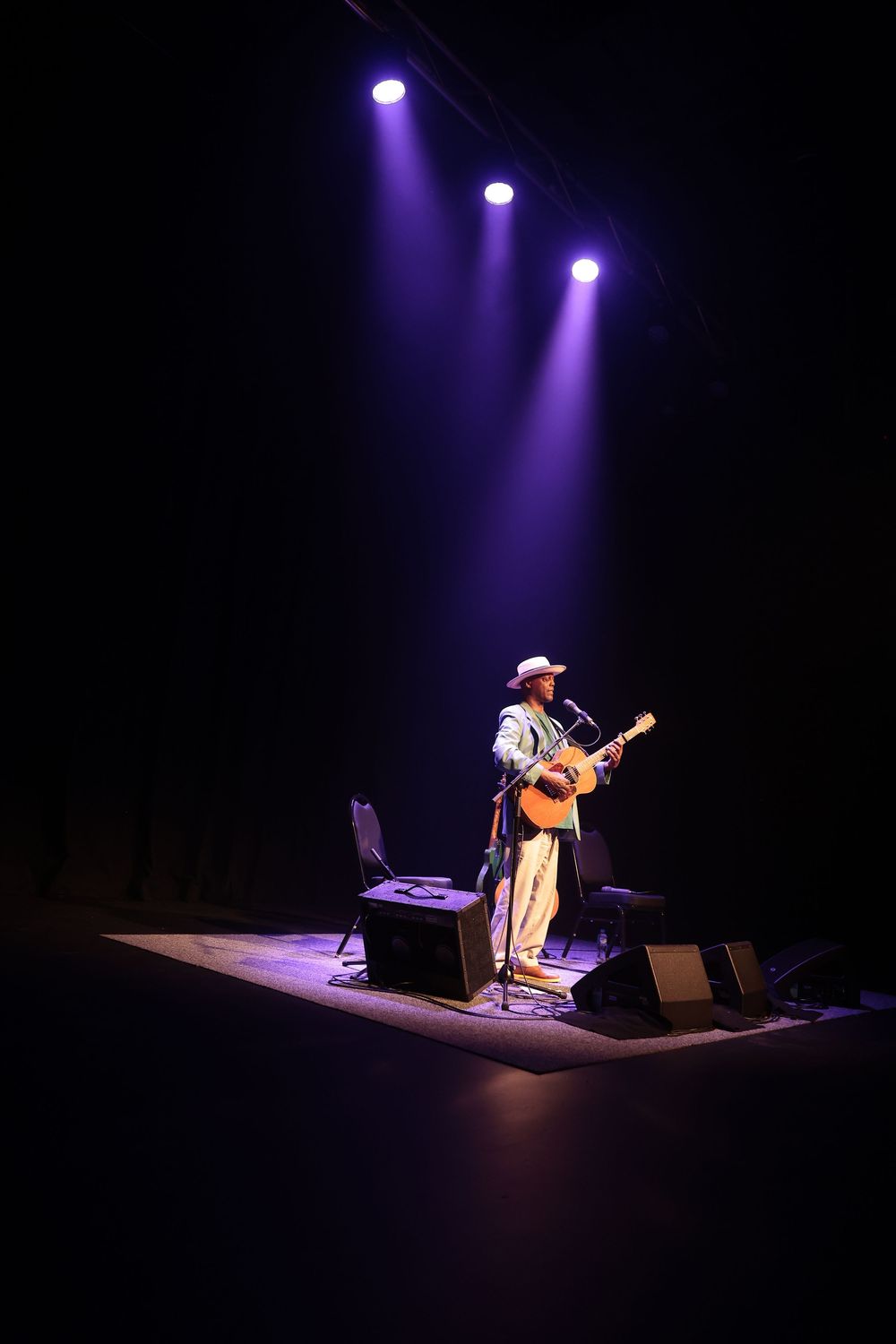 Solo musician in a white hat plays acoustic guitar under purple spotlights on a dark theater stage.