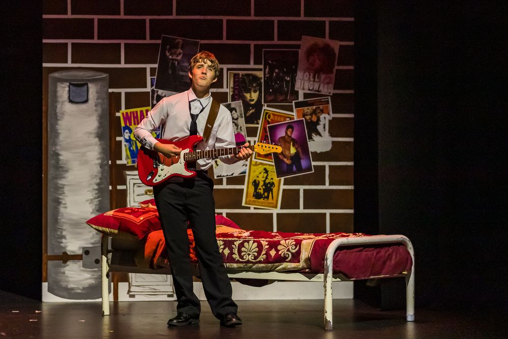 Young male actor plays electric guitar on a theater stage set designed to look like a teenage bedroom.
