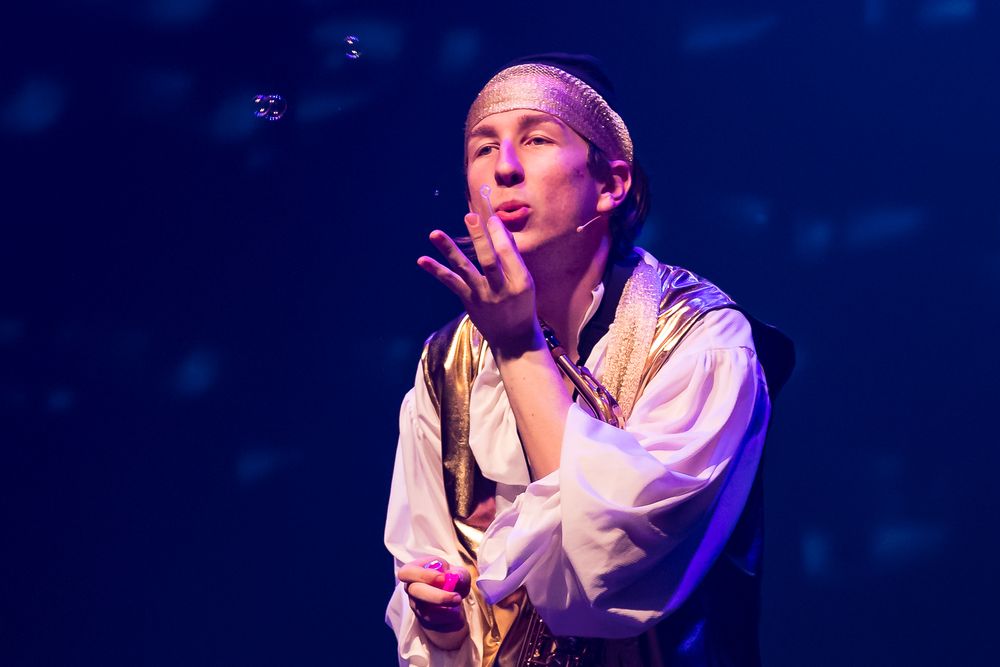 Young performer in gold and white costume blows bubbles on a blue-lit stage during a theater production.