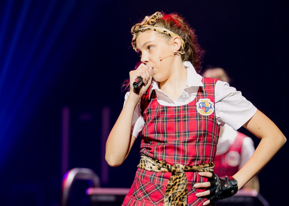 Young girl performs solo on stage in red tartan costume with microphone under spotlight.